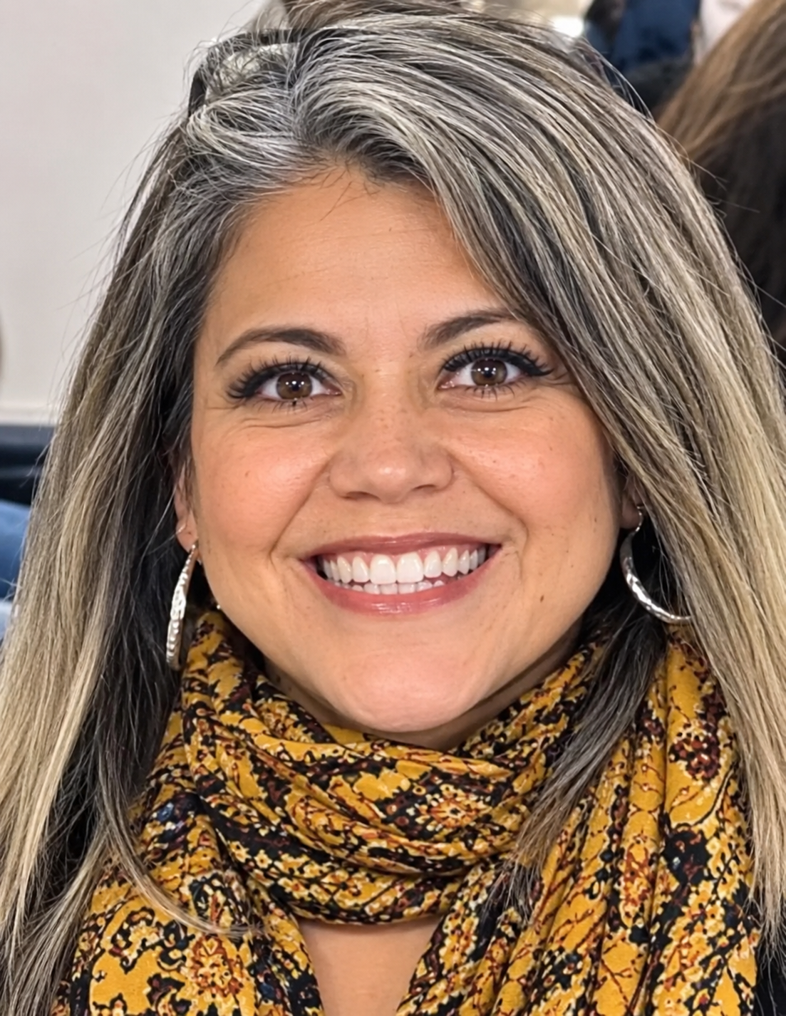 Portrait of a smiling woman with shoulder-length salt-and-pepper hair, wearing hoop earrings and a gold patterned scarf. She is looking directly at the camera indoors, with a softly blurred background.